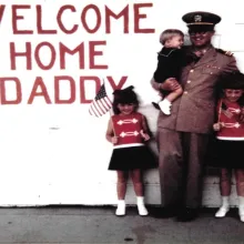 A man in a Navy uniform stands in front of a white garage door, that has "welcome home daddy" written on it in red. The man holds a small child, and two young girls are standing next to him holding American flags.