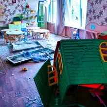 An elementary school classroom in Ukraine lies in ruins following a Russian bombing attack. The windows have been shattered, with glass littering the floor. Shards of glass are scattered across the children's furniture, creating a hazardous environment. In the foreground, a children's playhouse has also been destroyed, highlighting the extent of the devastation.