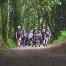A diverse group moves together along a forested path, sunlight filtering through moss-covered trees. At the center, a man in a wheelchair is pushed by a companion, surrounded by walkers of all abilities, embodying unity, resilience, and adventure.