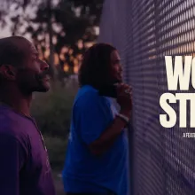 Two men standing at a fence, the first is in focus, his face looks stoic.