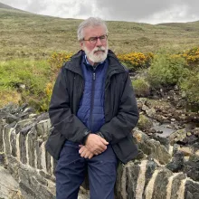 An older man with white hair and a beard, wearing glasses and a jacket, stands in front of a green field and hills. 