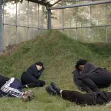 Four people are pictured, curled up in grass inside what appears to be a greenhouse. 