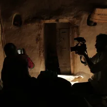 In an interior room, a man with a camera films a woman, who is also holding a camera.