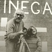 In front of a wall with "CINEGAEL" painted on it stands a father and his son, with a camera on a tripod.