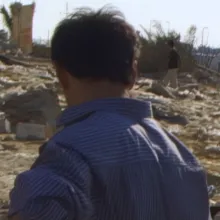 DV tape image of the back of a man's head, walking through a field with building rubble.