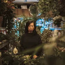 A woman with black hair stands in the middle of a plant shop.