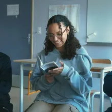 Three young people look at a book, while sitting in a classroom.