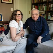 In a living room, a woman and a man wearing lavalier mics sit on a gray couch.
