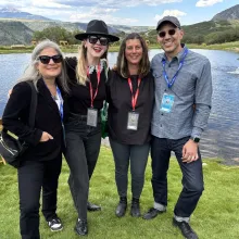 On the grassy banks of a lake, four white people wearing festival lanyards stand casually and happily.