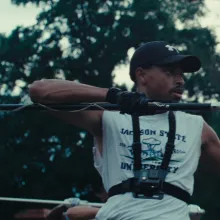 A drum major marches forward with a mace in hand. There are trees behind him.