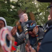 A group of powwow dancers outside, in front of a copse of trees.