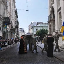 Led by a soldier carrying the Ukrainian flag, a casket is being carried by soldiers into a church; some onlookers kneel on the street as if it were a parade.