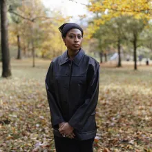 A Black woman in a black leather jacket stands with hands clasped in a field of autumnal leaves.
