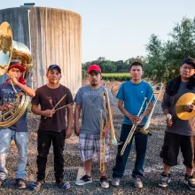 Five young men, each holding a different musical instrument, stand outside in a sunny field