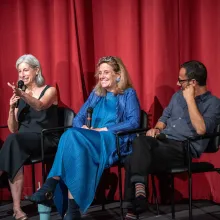 In front of red curtains, three people sit on chairs on a stage.