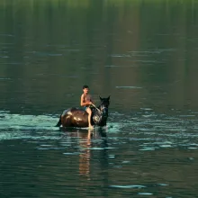 A drenched young man in nothing but a swimsuit rides a horse in the middle of a body of water