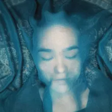 Under a blue cloth that is semi-transparent, a teenaged girl lays on the floor with her eyes closed.