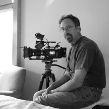 Black and white image of a white man with short hair, glasses and goatee, man sitting in a room next to a camera, as another stands by a window.