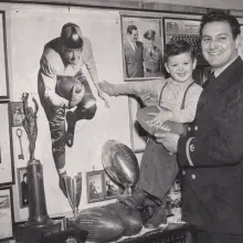 Black and white photo of Sid holding his son Bobby. They are in front of a poster of Bobby as a quarterback.