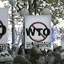 Grainy shot of group of outdoor protestors holding "We Demand Public Voice" signs with WTO crossed out
