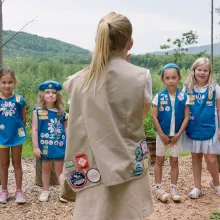 Out in a lush green forest, six young girl scouts in blue uniforms look at an older blond girl scout in a tan uniform whose back is against the camera