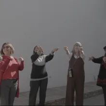 Seven women in their 70s and 80s stand in a semi-circle doing stretches in a courtyard bathed in sunlight.