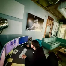 A white man with long black hair in a hoodie sits at an editing bay in a stylish, industrial-looking, emerald-decorated room
