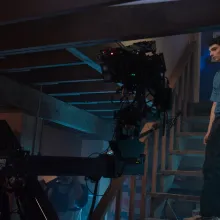 A young white actor wearing a blue button down shirt and pants stands on a basement with a camera rigged in front of him