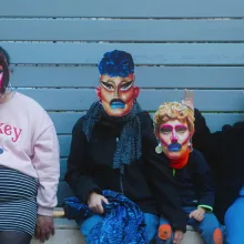 Four people sit against a light blue wall wearing regular clothes and paper masks of drag queens