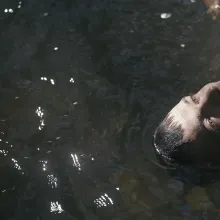 A white man with dark hair and hazel eyes looks up as he floats on a body of water