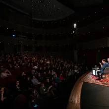 At a stage, two men sit in chairs as a rapt theater audience looks on