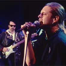 A man with a blond ponytail sings passionately into a microphone, in front of the rest of his rock band.