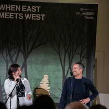 A light-skinned woman with dark hair and glasses, wearing a black shirt and a white cardigan talks into a microphone while talking to a white man in a black sweater with short-cropped hair with a poster advertising When East Meets West