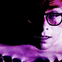 A magenta-tinted image of a man with glasses, leaning forward.