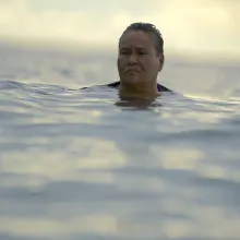 A brown, heavy-set woman's head is seen above ocean water against a sunny sky behind her