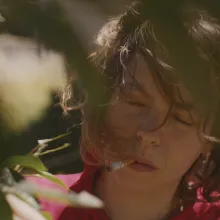 A close up of a white woman with shoulder-length dark hair in a red shirt, eyes closed, holding a cigarette in her mouth outside