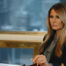 A heavily-made up white woman with long, colored blond hair sits a desk with a stern look on her face