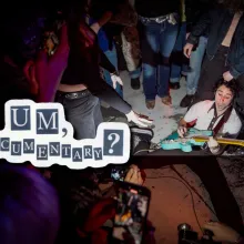 Eli, the frontman of Um, Jennifer?, is on the ground playing a blue guitar. He is surrounded by a swarm of energetic fans recording and cheering him on. The image is captured from an Um, Jennifer? show in Brooklyn. He is wearing gray trousers and a white blouse.