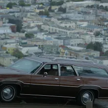 A brown station wagon is parked atop a street that overlooks a city down below; an older woman can be seen sitting in the backseat while a man stands nearby in the foreground