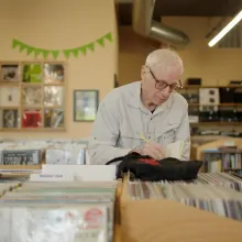 An elderly white man with thin white hair and glasses jots something on a notebook while in the middle of an old-school record store