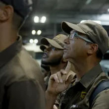 A group of men dressed in brown UPS uniforms look up, while standing inside a warehouse.
