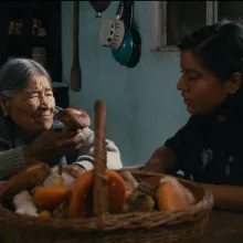 An elder Indigenous woman with long grey hair parted and pulled back holds a mushroom while sitting at a table with a younger Indigenous woman with dark black hair, a basket full of mushrooms in front of them both.