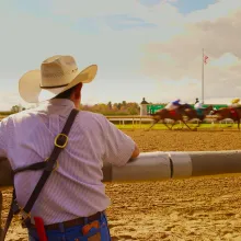 A man in a cowbow hat seen from behind is watching a horse race happening across from him