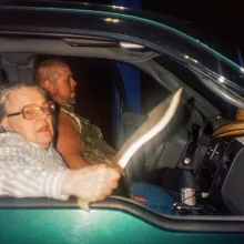 An older women with glasses sitting in a truck at night wields a knife at the camera