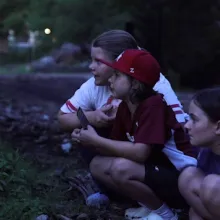 Three children at dusk in the woods crouched down looking into the trees and grass