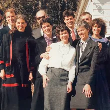 A group of ten siblings stand outside against a white house in late 1980s formal attire. They smile and squint in the sun. Their father, elderly, stands in the back and is mostly obscured.