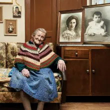 An elderly woman with pulled back grey hair sits on a yellow-print heavy coach surrounded by framed black and white photos of her younger self 
