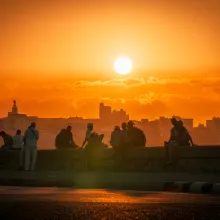 Silhouettes of people sitting and standing along the Malecón seawall in Havana at sunset, with the city skyline behind them and a deep orange sky.