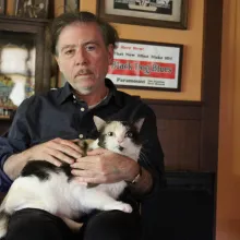 An older white man with thinning black hair and a bit of stubble, wears a dark blue shirt and dark pants as he sits staring at the camera as a white and black cat sits on his lap