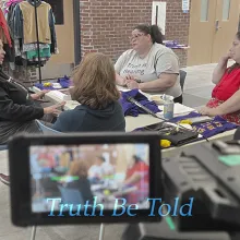 Seen through a camera's viewfinder, four women gather around a table — one wearing a shirt that reads "Truth is Healing" — as Truth Be Told documents stories Vermont has never officially heard before.